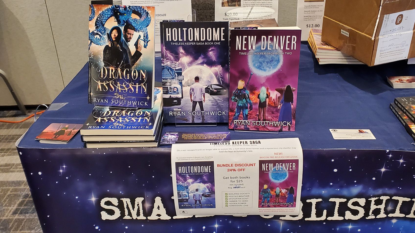 3 books standing up on a table: Dragon Assassin, Holtondome, New Denver. A discount sign in front.