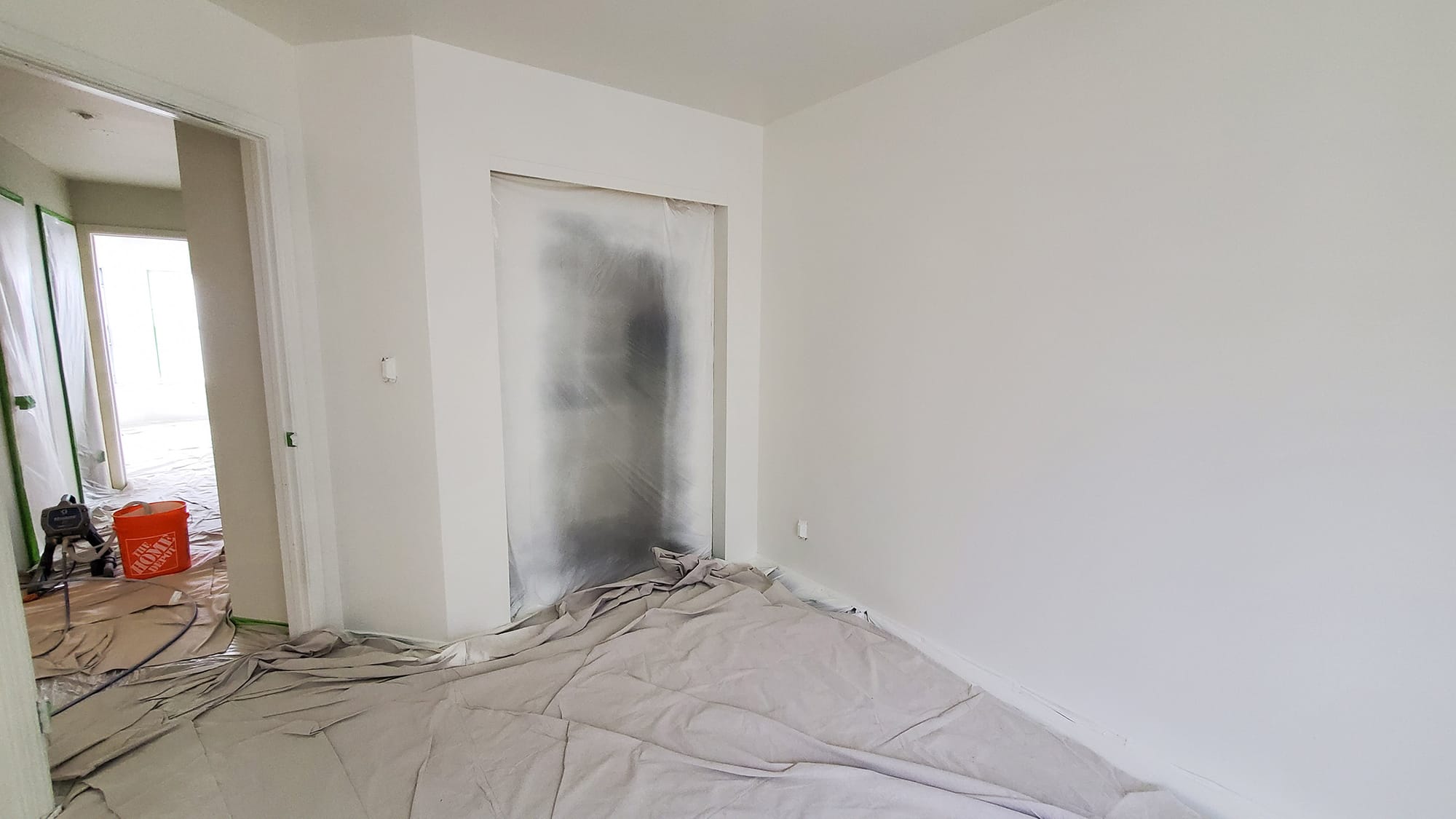 White-walled bedroom with canvas on the floor, plastic drape over the closet, and a paint sprayer in the hall