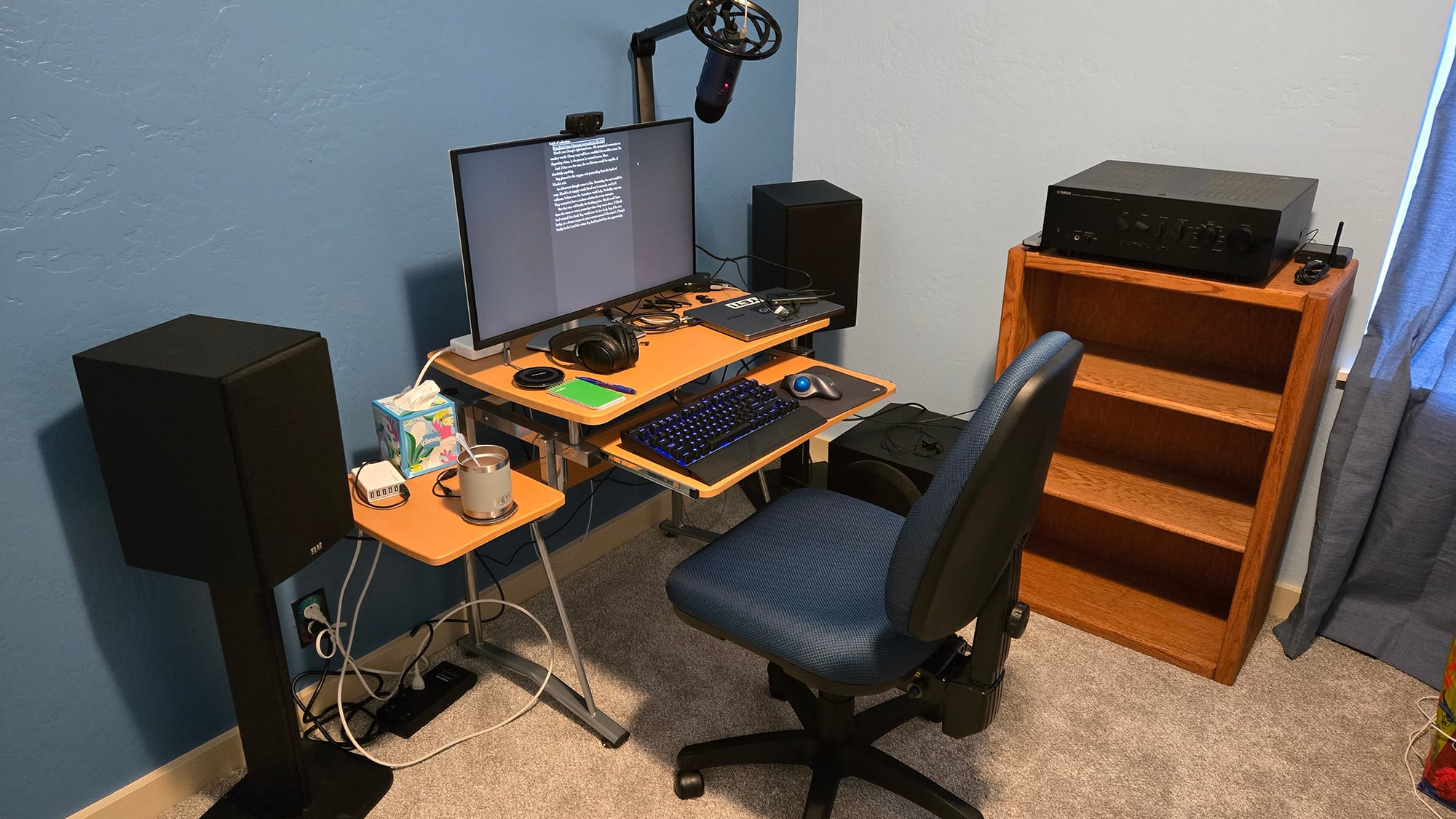 Small computer desk against a blue wall. Speakers and a stereo on an empty bookshelf. Coffee. A boom mic.