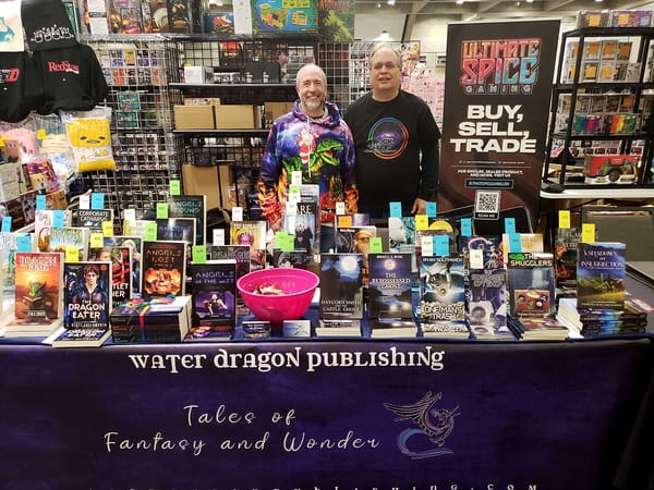 Two men standing behind a table full of books at a comic convention. Banner: "Water Dragon Publishing"